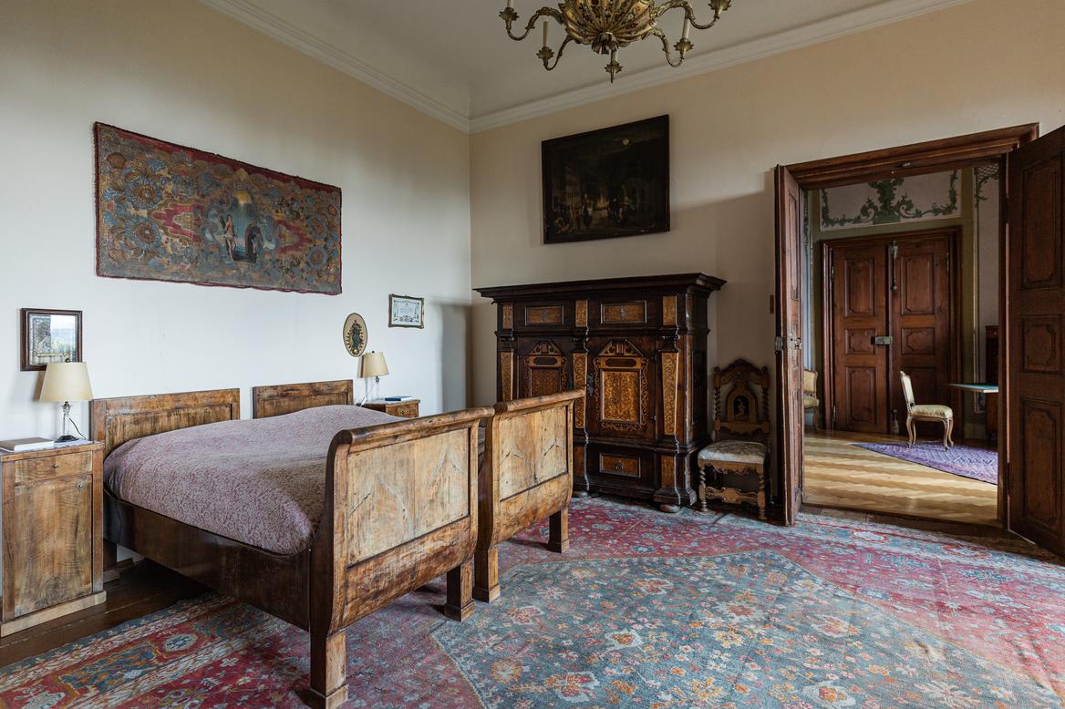Zwei Holzbetten unter einem Wandteppich, dahinter der Blick durch eine geöffnete Türe ins Wohnzimmer